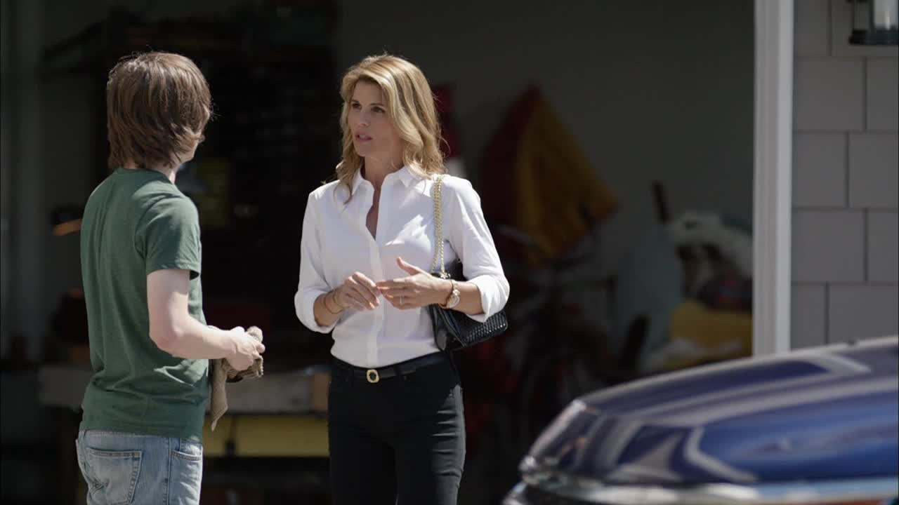 A woman in a white shirt and black pants gestures with her hands as she speaks to a young man, who holds a cloth in his hands. They're standing in front of a garage, the sunlight bright on their faces.
