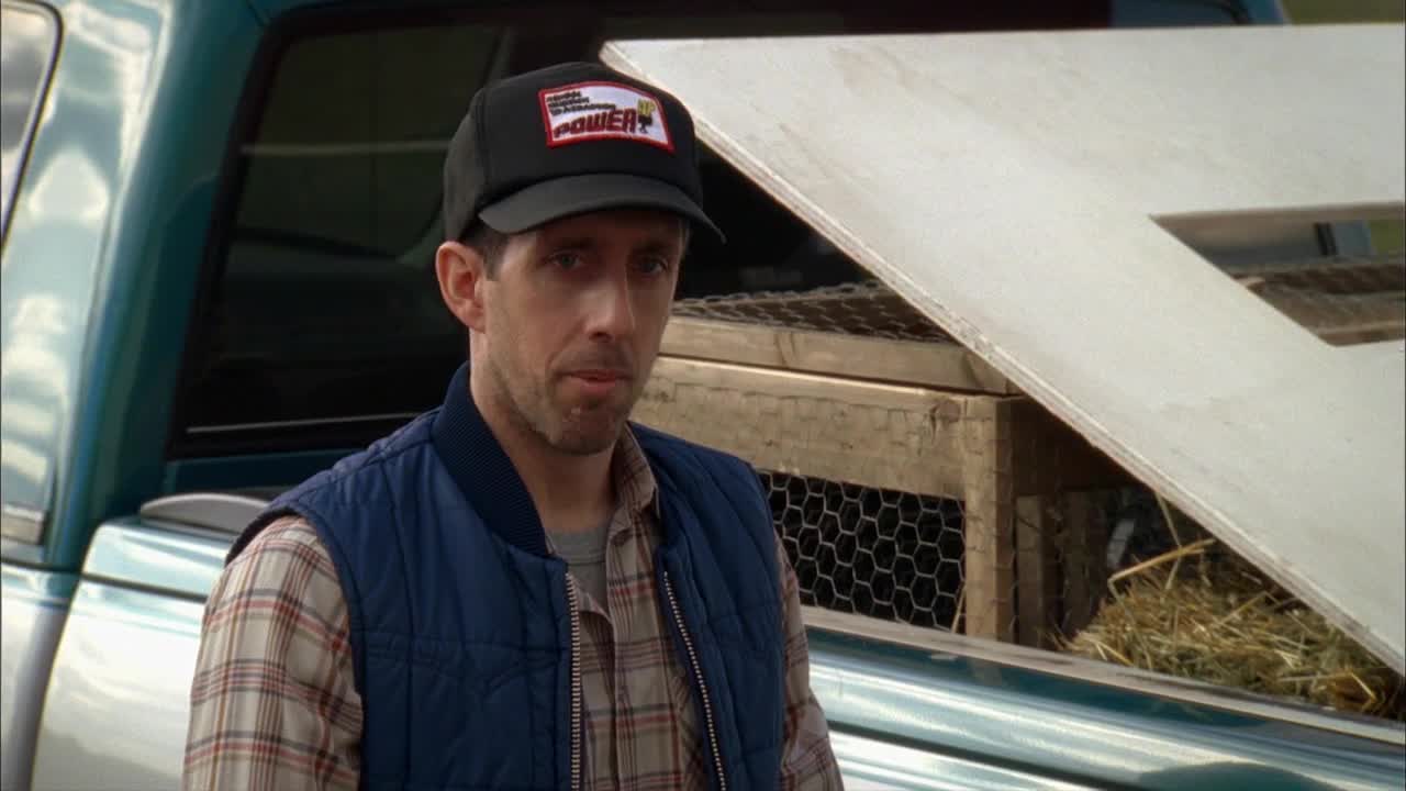The man in the black hat is looking straight ahead, standing next to the truck bed. A chicken coop sits in the back, with a large piece of plywood leaning against it.
