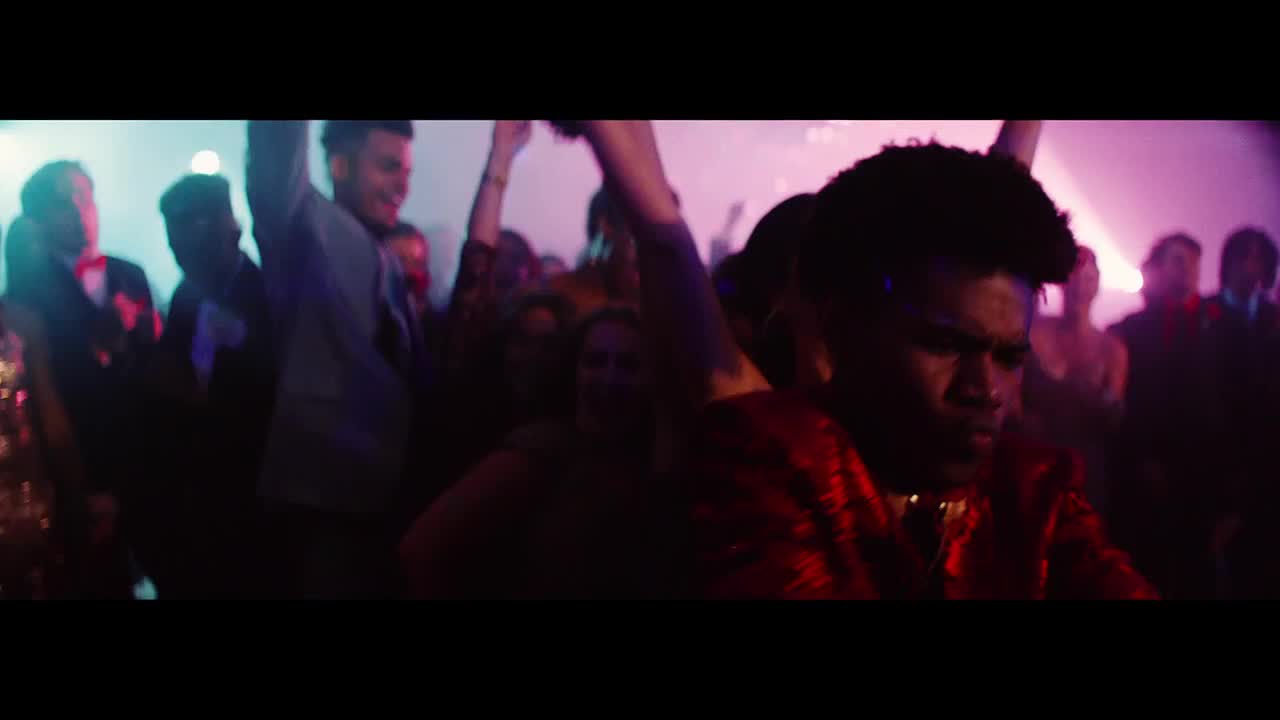 A crowd of people are dancing with their arms in the air, bathed in purple and blue light. The person in the foreground, wearing a red jacket, seems to be focused on the music.
