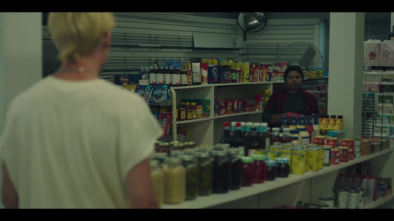 A customer with bleached-blonde hair stands at the counter of a convenience store, facing the cashier. Rows of jars and bottles line the shelves, and the cashier stands ready to assist.
