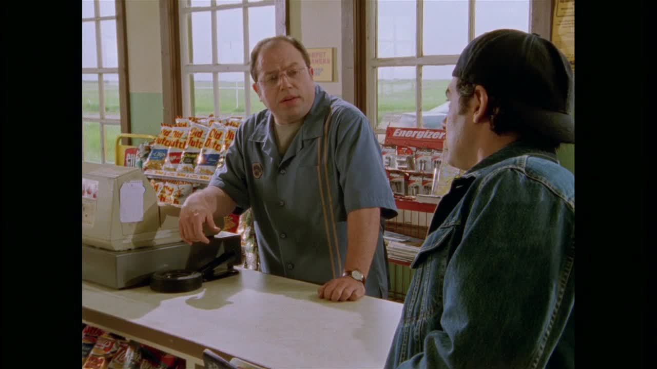 The man behind the counter, wearing a blue shirt with suspenders, is gesturing with his hands as he talks to the man in the denim jacket.  They're standing in a small store, with shelves of snacks and magazines visible behind them.
