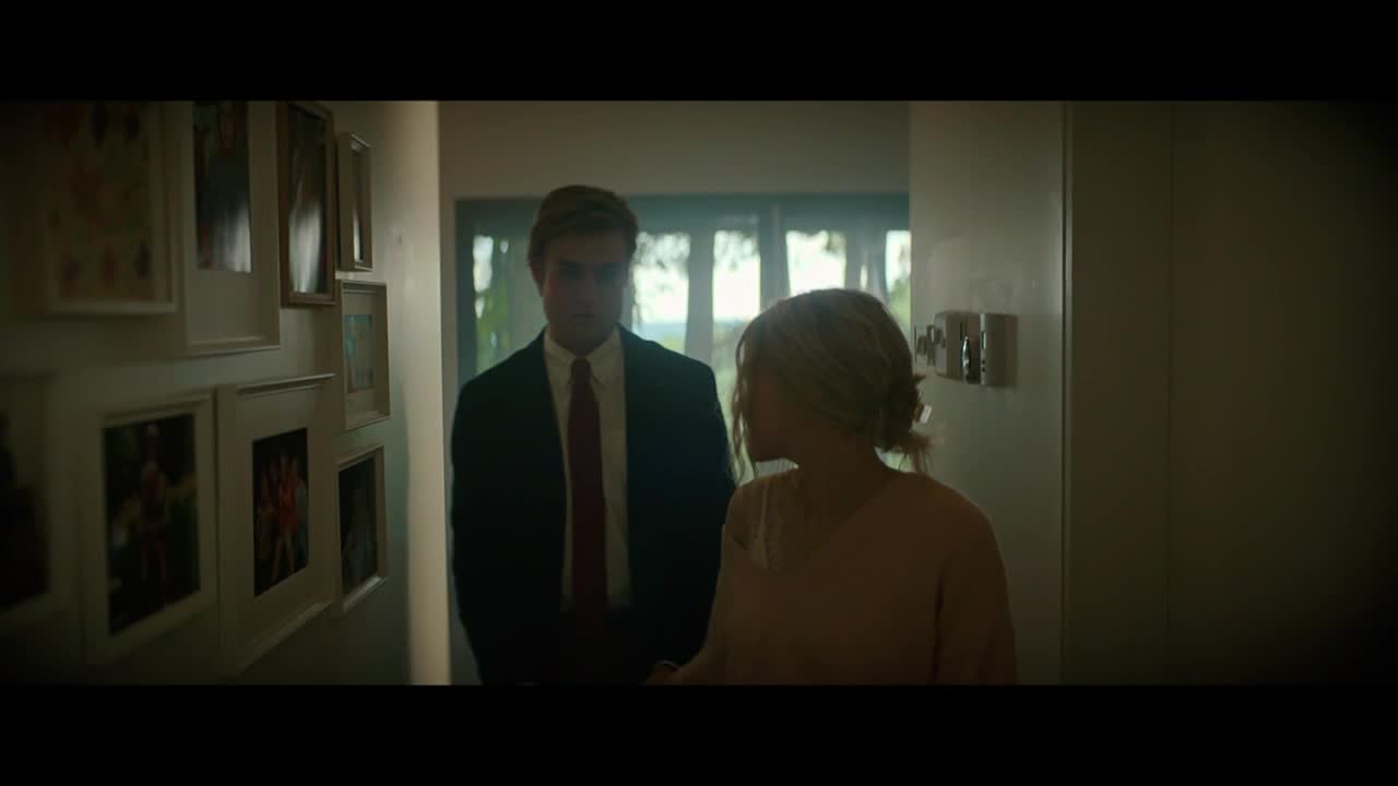 A man in a suit and red tie stands in a hallway, looking at a woman with blonde hair. She faces away from the camera, and they seem to be having a serious conversation.
