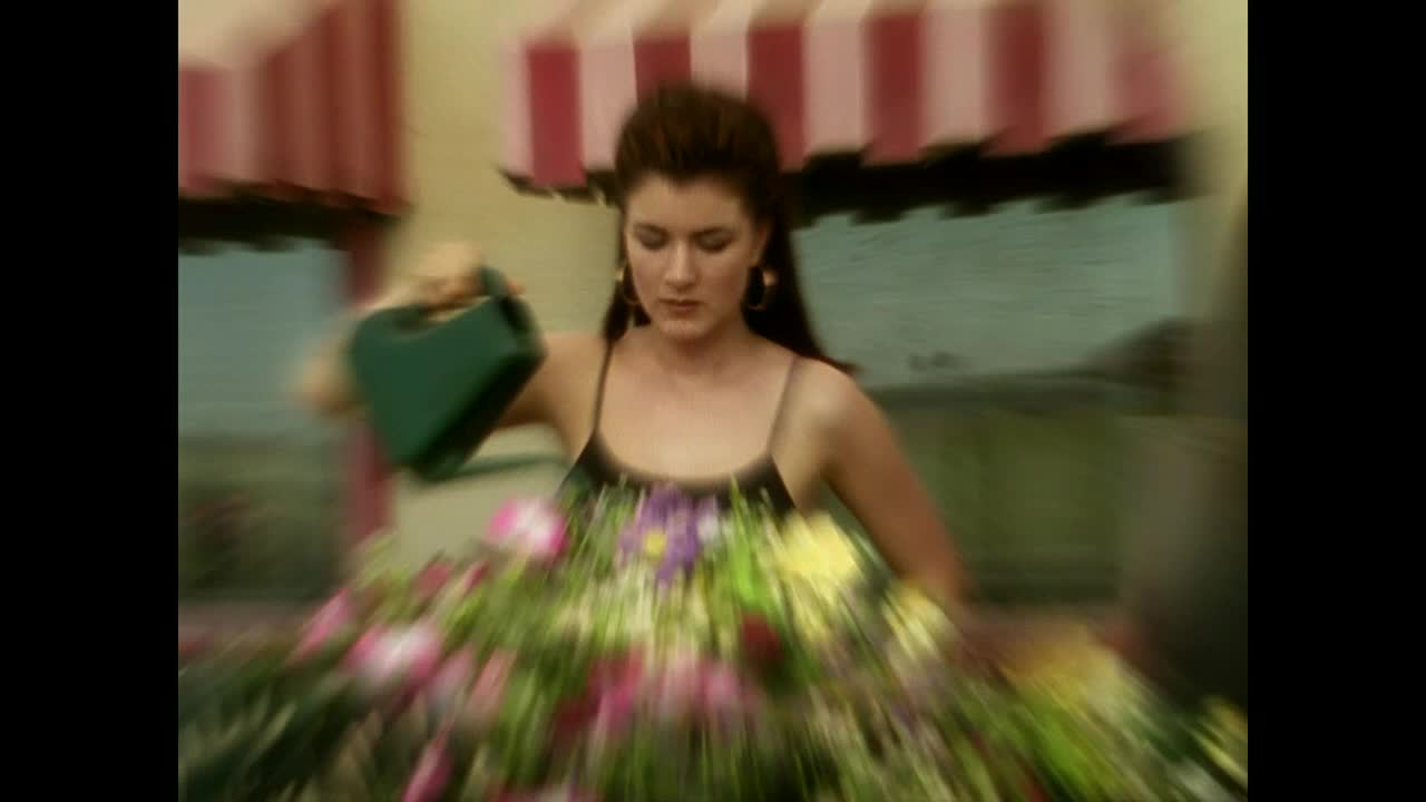 A woman in a black tank top leans over a vibrant flower box, holding a green watering can. The flowers are a mix of pink, purple, and yellow, and the whole scene has a soft, slightly blurred quality.
