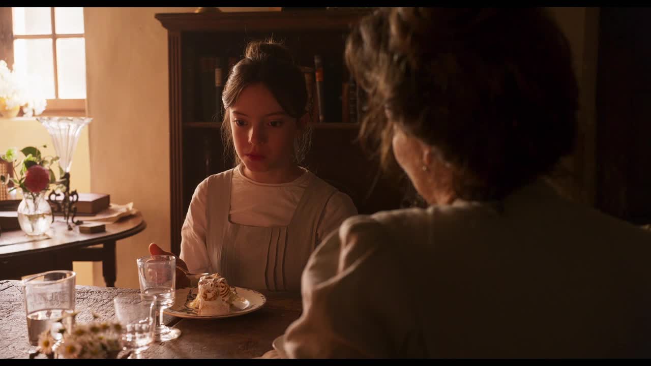 A young woman in a light-colored dress sits at a table, looking down at a plate with a dessert. Across from her, a woman with dark hair is partially visible, also at the table, in what looks like a scene from a period drama on Crave 4 in Canada.
