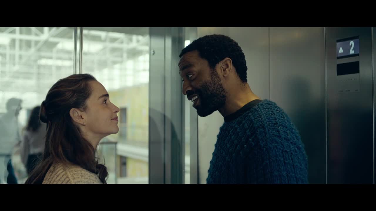 Inside an elevator, a man in a blue sweater is speaking, his eyes crinkling with a smile, as he looks at a woman. The woman, with her hair pulled back, gazes up at him, her expression thoughtful.
