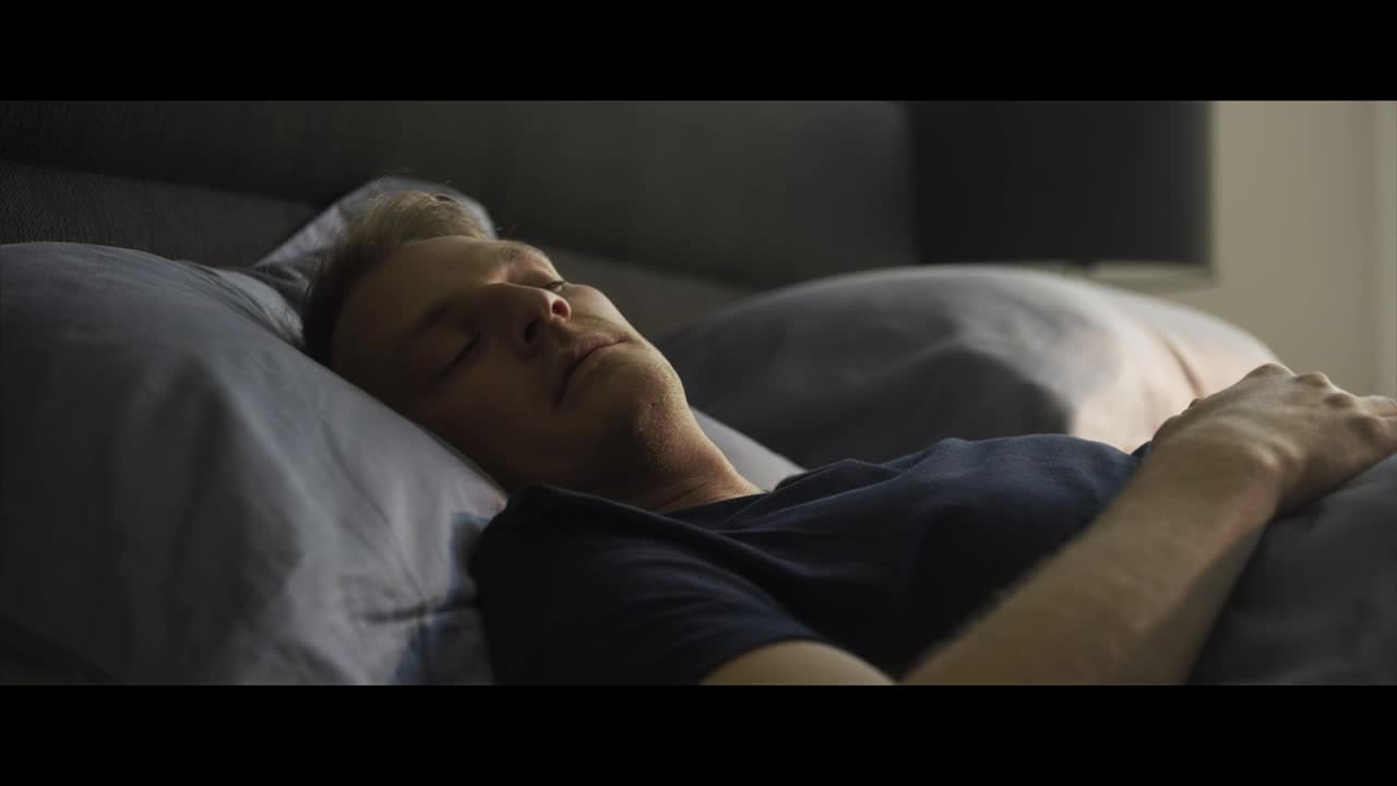 A man lies asleep on a pillow, his face illuminated by soft light. His hand rests on the bed, still and relaxed.
