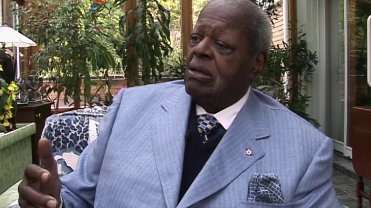 The man in the light blue suit gestures with his right hand as he speaks, his expression serious. He's seated in what appears to be a sunroom, surrounded by plants and natural light.
