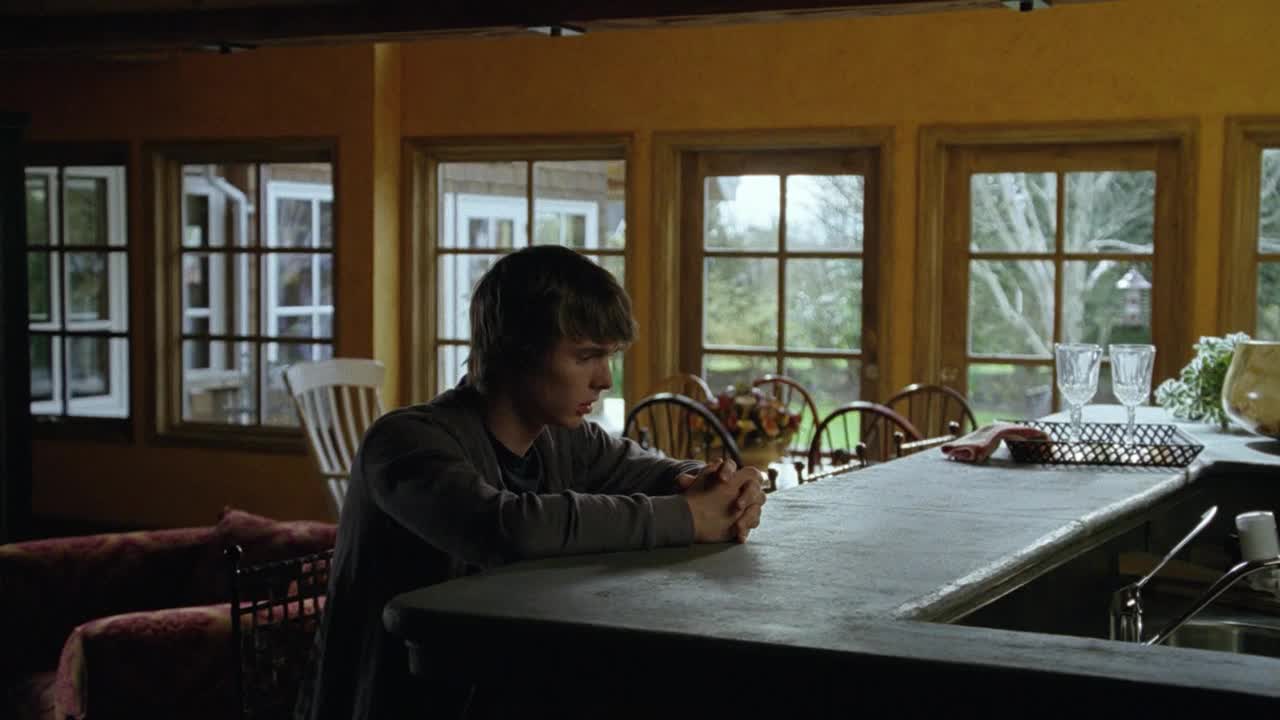 A young man sits at a kitchen counter, his hands clasped and head bowed. Outside the windows, the trees are bare, and the light is dim.
