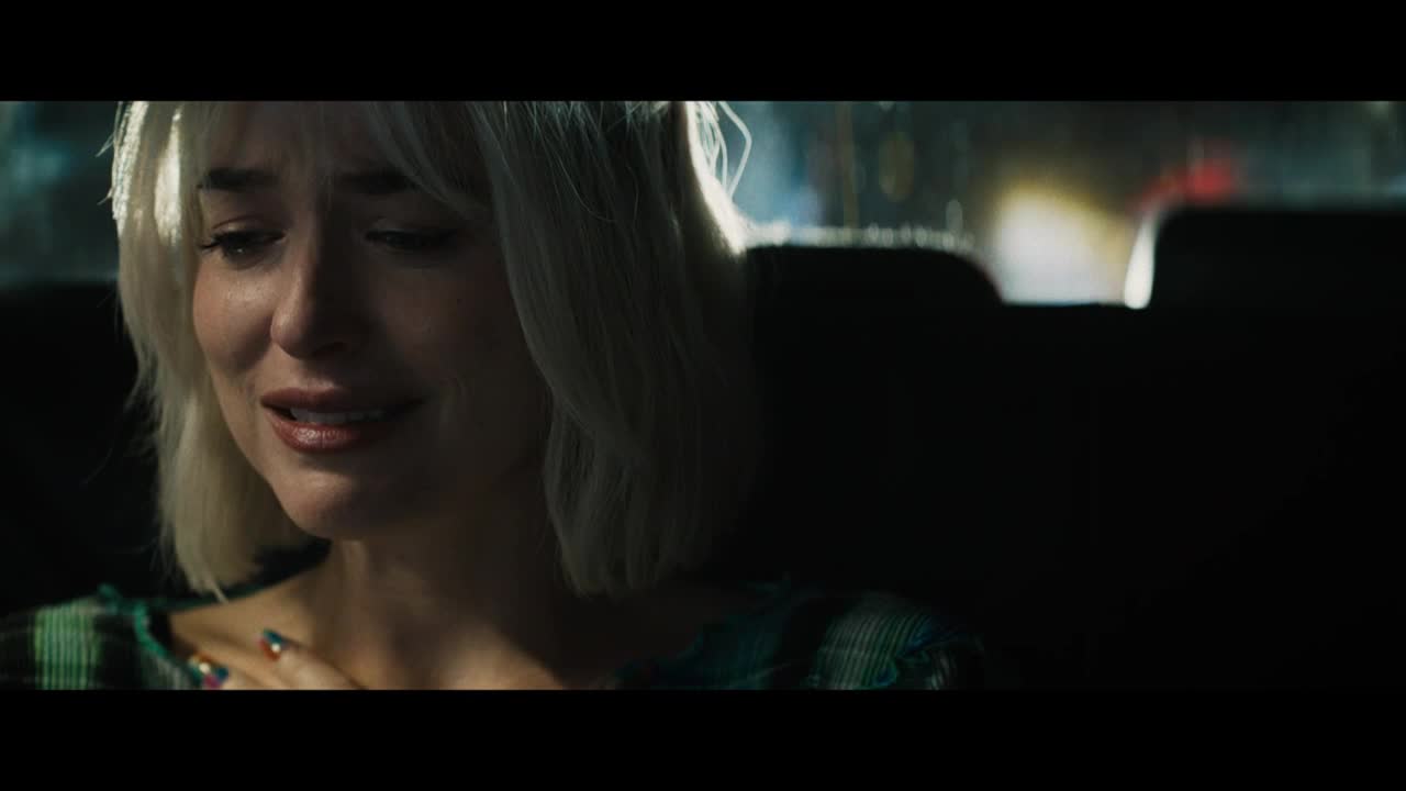 Tears stream down her face as she sits in the back of a car, her hand resting on her chest. The light from outside reflects in her eyes, highlighting her distress.
