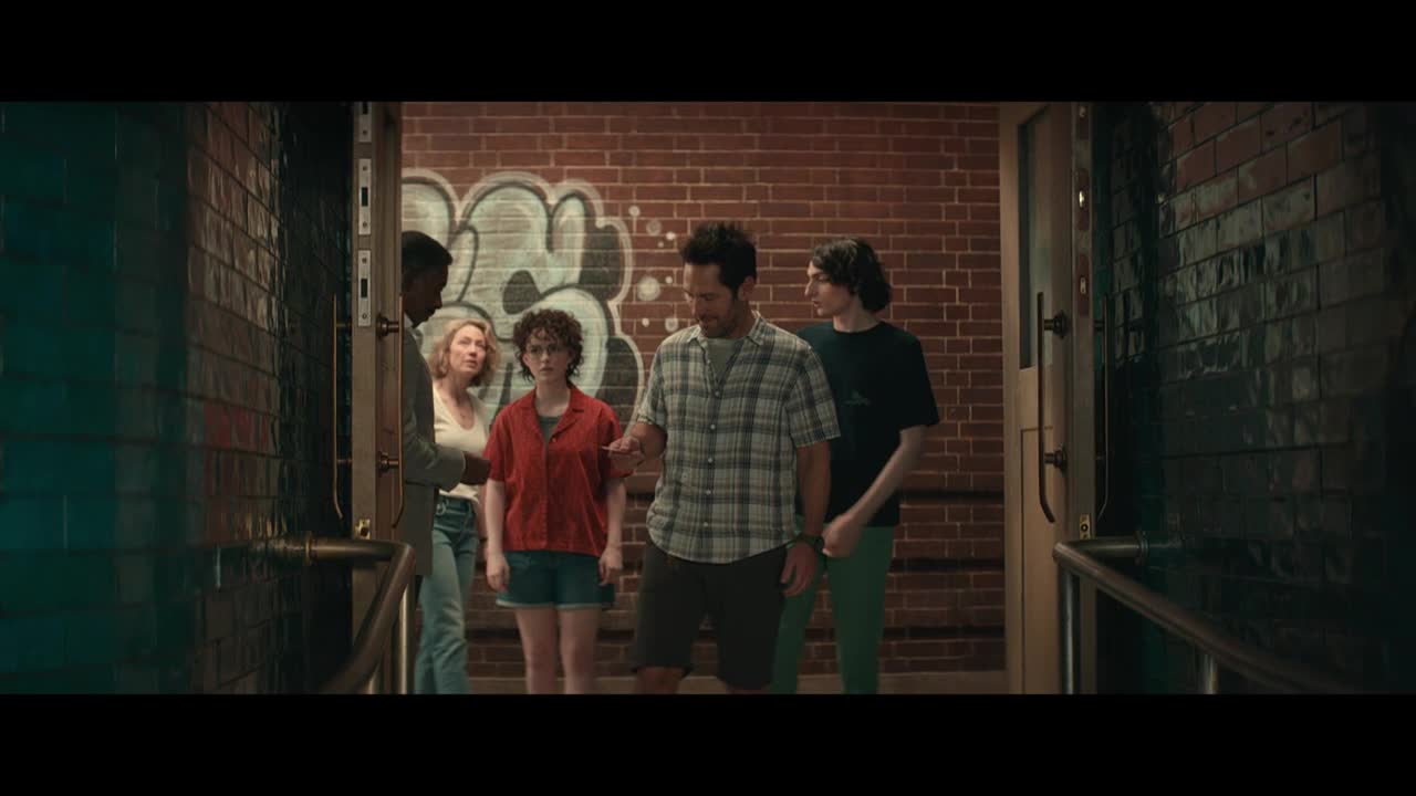 A group of people stand in a brick-lined hallway, framed by a doorway with graffiti on the far wall. The man in the plaid shirt gestures toward a piece of paper in his hand, while the others look on.
