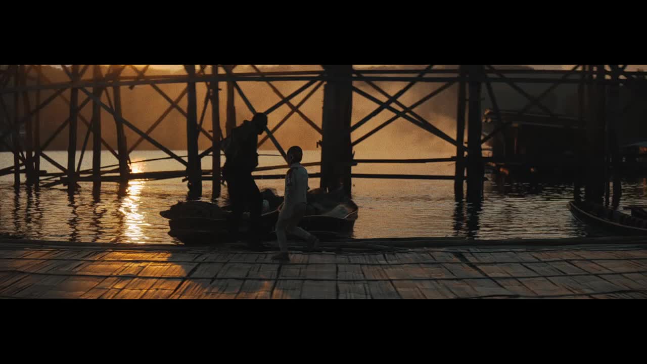 A man helps a boy out of a small boat onto the wooden dock. The sun reflects off the water, casting long shadows from the pier's supports.
