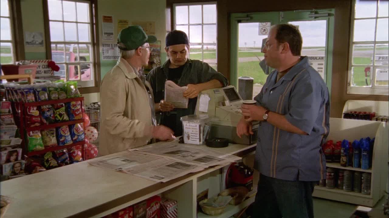 The guy in the green cap gestures while the younger man unfolds a newspaper at the counter. A man in a blue shirt holds a coffee cup and watches the scene unfold.
