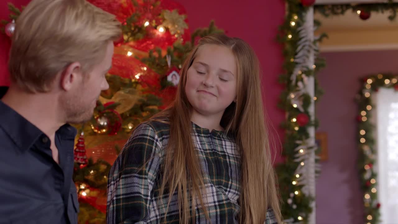 A man in a dark shirt looks at a girl in a plaid top, who's smiling with her eyes closed. Behind them, a decorated Christmas tree and garland frame the doorway.
