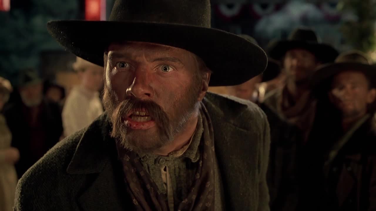 The man in the black hat stares straight ahead, his face streaked with grime and a cut on his lip. Behind him, other men in hats and coats stand in a crowd, watching something off-screen.

