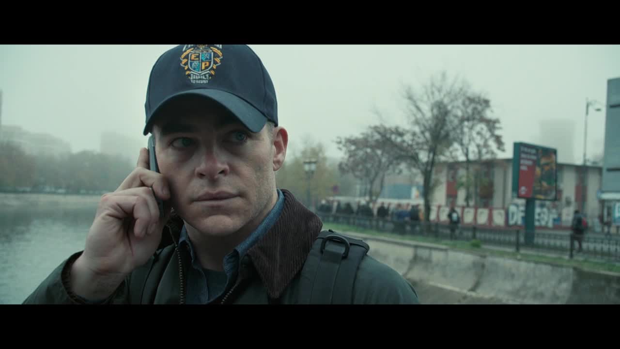 A man in a cap, with a patch, holds a phone to his ear, his gaze fixed on something off-camera. Behind him, a few people mill about near a body of water, and a billboard advertises something.
