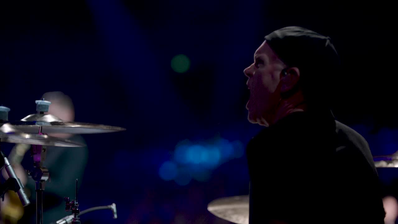 The drummer, in a black beanie, is mid-song, his mouth open as he plays. The cymbals shimmer in the stage lights, reflecting the blue and green hues of the background.

