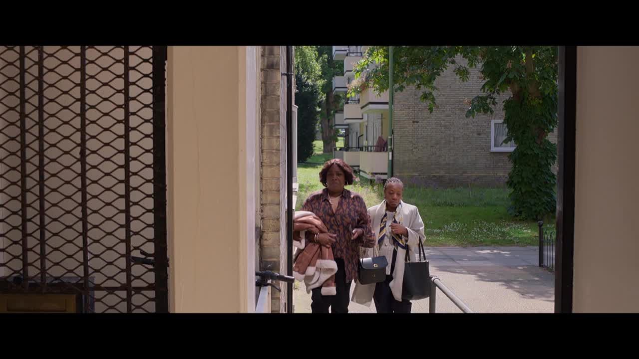 Two women walk towards the camera, emerging from a gated entrance. One carries a blanket, the other a purse and a bag.
