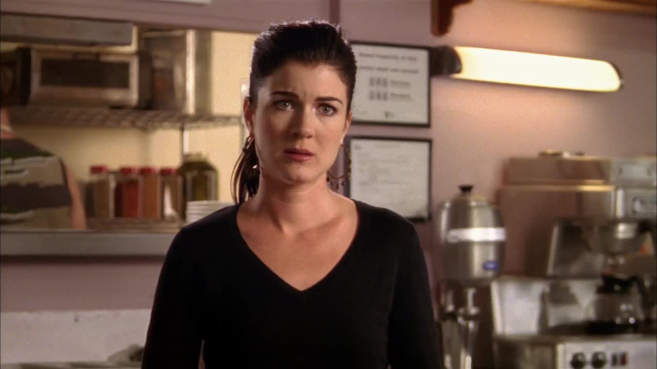 The woman in the black top stares directly ahead, her expression a mix of concern and surprise. Behind her, the kitchen is lit by a long fluorescent light, and the scene feels like a moment from a Canadian drama.
