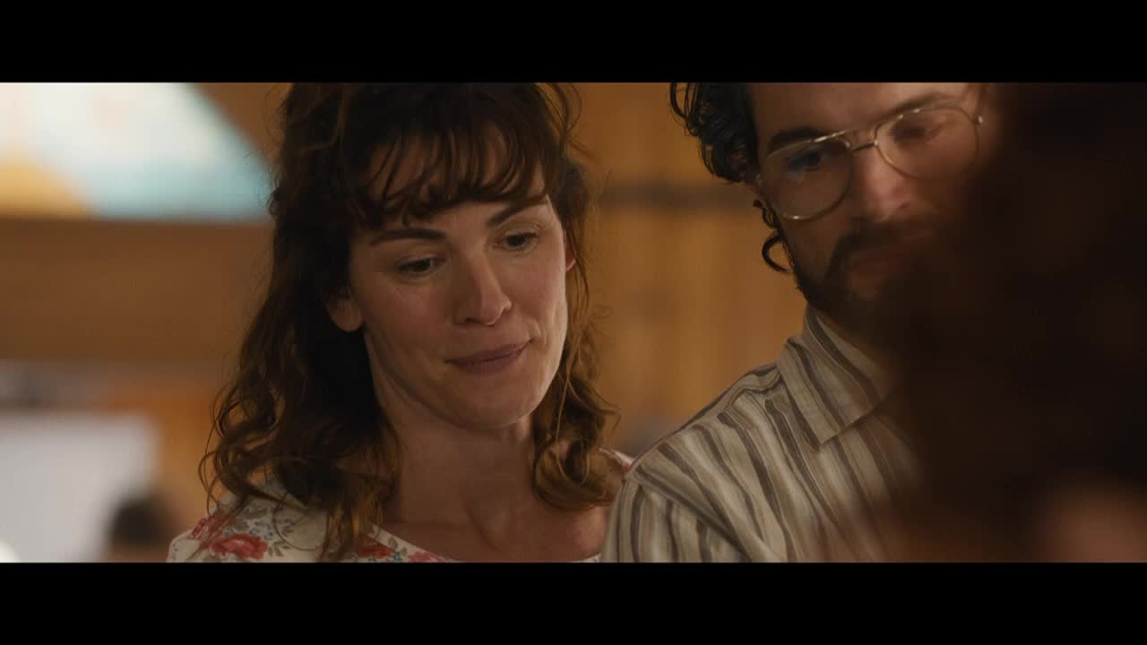 A woman with curly brown hair smiles softly, her gaze lowered. Beside her, a man in glasses looks down, a serious expression on his face.

