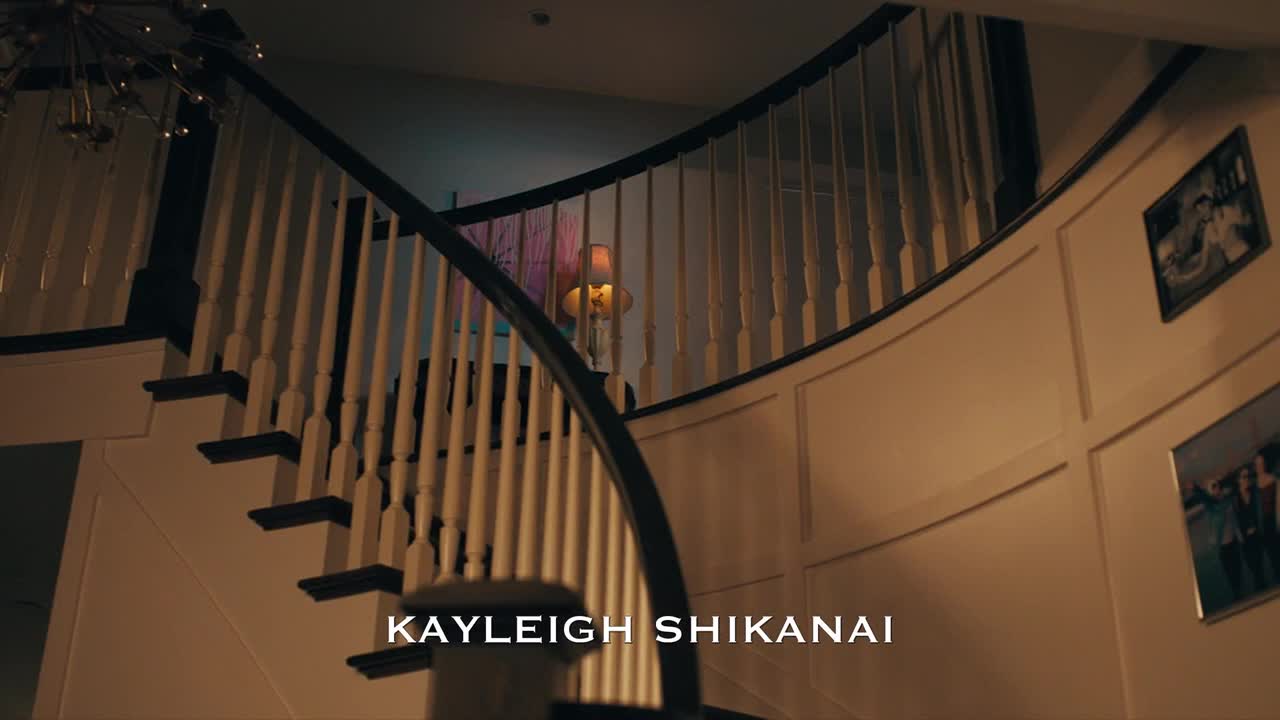 A curved staircase, framed by a dark railing and white spindles, sweeps upward. A lamp glows softly on the landing, and the name "Kayleigh Shikanai" appears at the bottom of the screen, likely from a show on Crave 4 in Canada.

