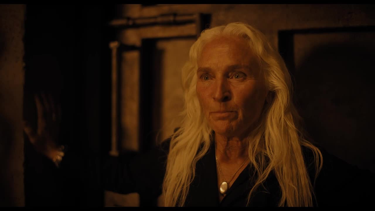 A woman with long, white hair and a serious expression stands in a dimly lit space. The light catches her face, and she's wearing a pendant.
