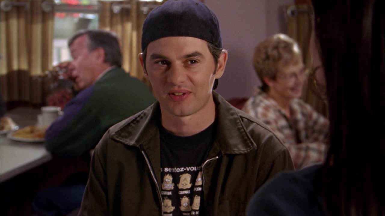 The guy in the cap is talking, looking right at someone off-screen, while a couple of other people sit in the background. The diner scene feels familiar, like something you'd see on Crave 4.
