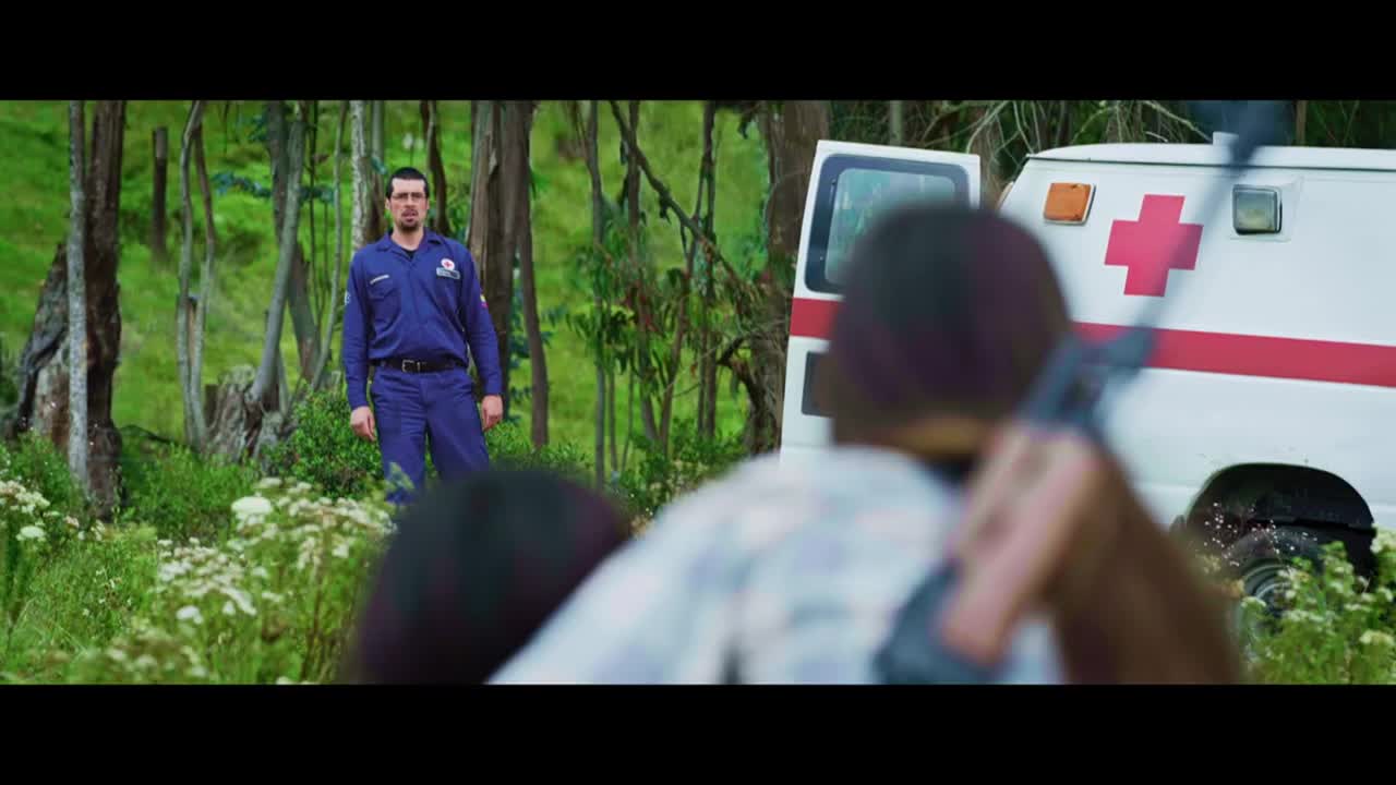 A man in a blue uniform stands in a clearing, watching. The back of an ambulance is visible, and someone is aiming a gun in its direction.
