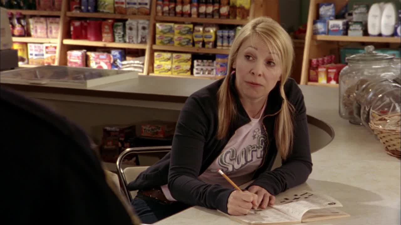 A woman with blonde pigtails leans over a notepad, a pencil in her hand. Behind her, shelves are stocked with snacks and drinks, the setting of a corner store.
