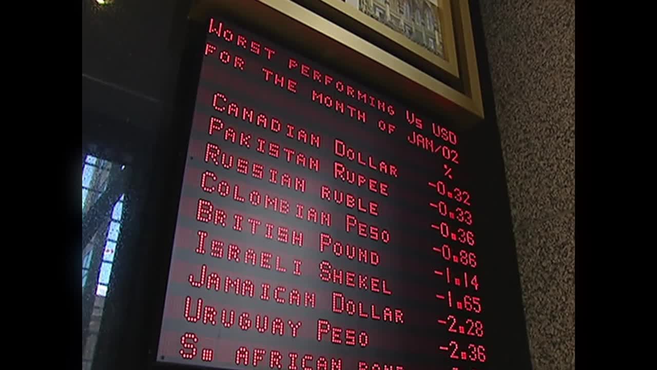 A red LED display shows currency performance, and the Canadian dollar is listed near the top, down 0.32% against the US dollar in January 2002. The screen is framed in dark wood, likely in a financial setting, possibly on a Canadian news broadcast like Crave 4.
