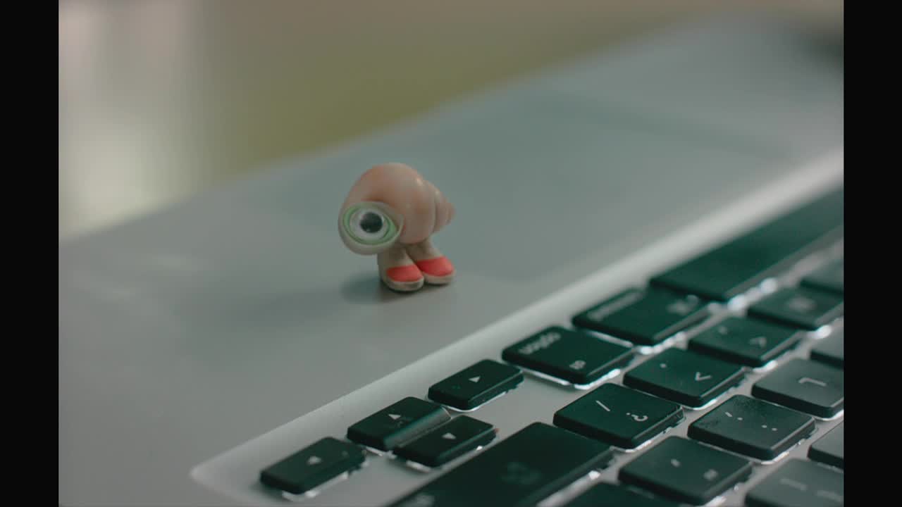A tiny, cartoonish snail with red shoes stands on a laptop's silver surface. Its big, spiraled eye is focused on something beyond the frame.

