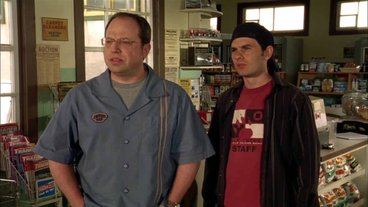 Two guys are standing inside a small shop, looking off to the side. The man in the blue work shirt seems concerned, while the other in the red shirt and black bandana appears to be listening intently.
