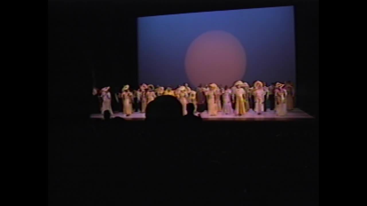 A group of figures in long robes and wide-brimmed hats stand on a stage, bathed in dim light. Behind them, a large, pale circle glows against a dark blue backdrop.
