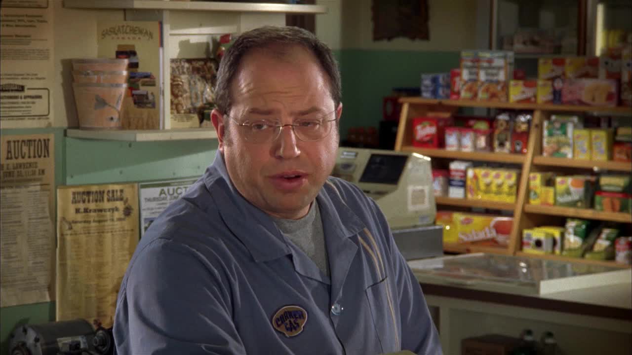 Brent Leroy, from Corner Gas, is behind the counter, talking. He's wearing his work shirt, and the shelves behind him are stocked with snacks.
