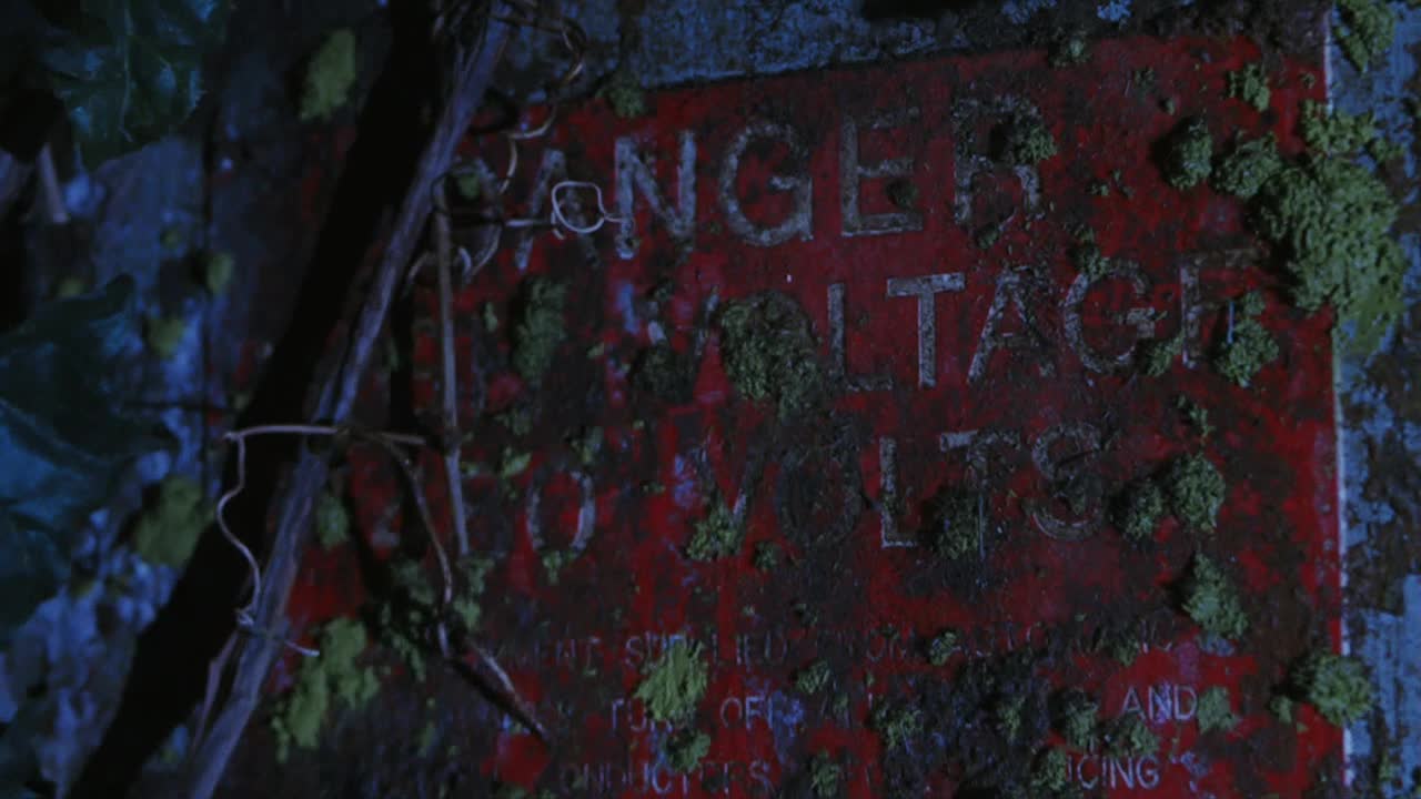 A weathered red sign warns of high voltage, the lettering barely visible beneath a layer of moss.  Twisted wire and vines partially obscure the sign, giving it a forgotten, ominous feel.
