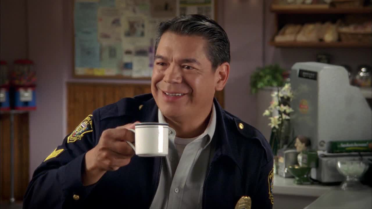 The police officer, from a Canadian show on Crave 4, is smiling as he holds a white mug, taking a moment to enjoy a cup of coffee. He's in a diner, the backdrop filled with the usual coffee shop clutter.

