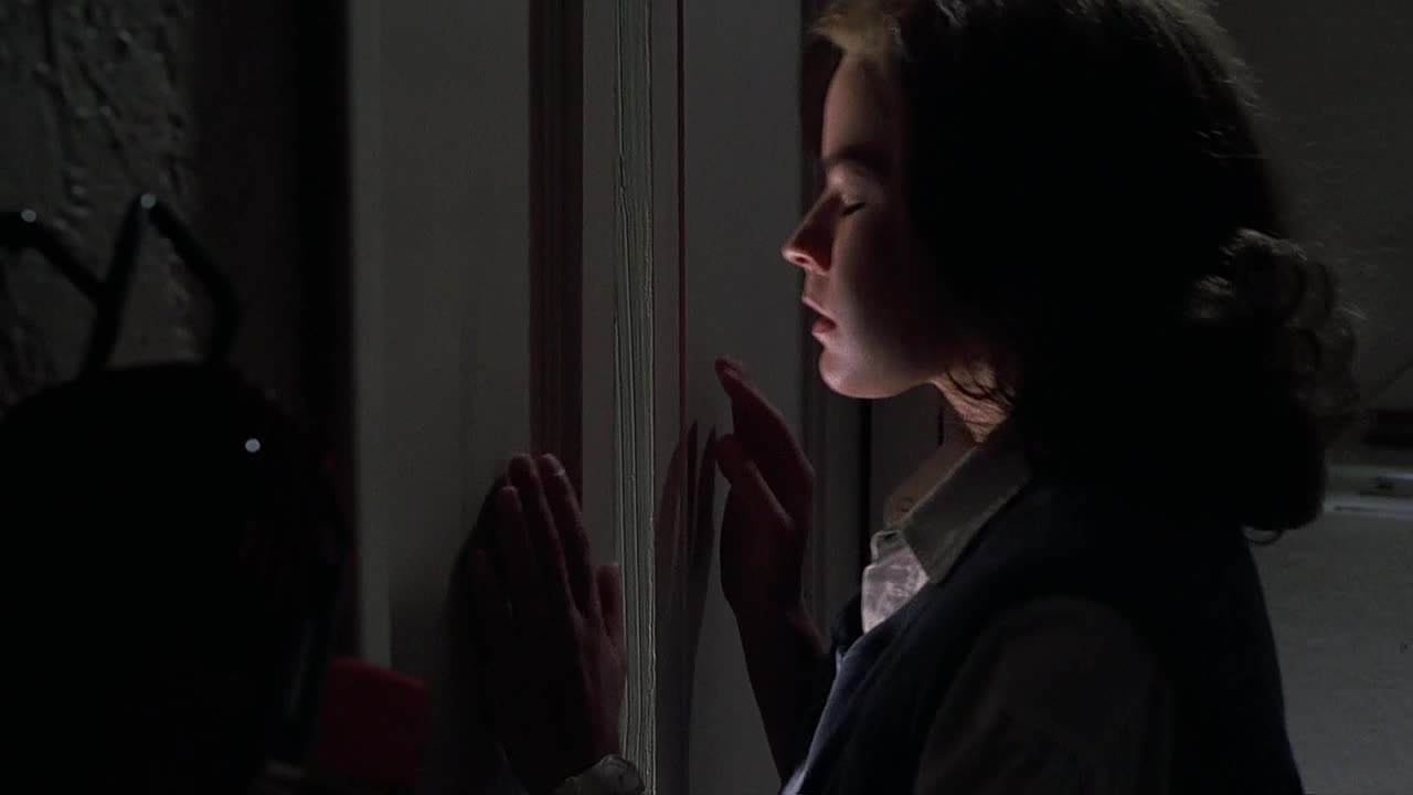 A woman, eyes closed, presses her hands against a door frame, her face illuminated by a sliver of light. The darkness obscures the room beyond, but the tension is palpable.
