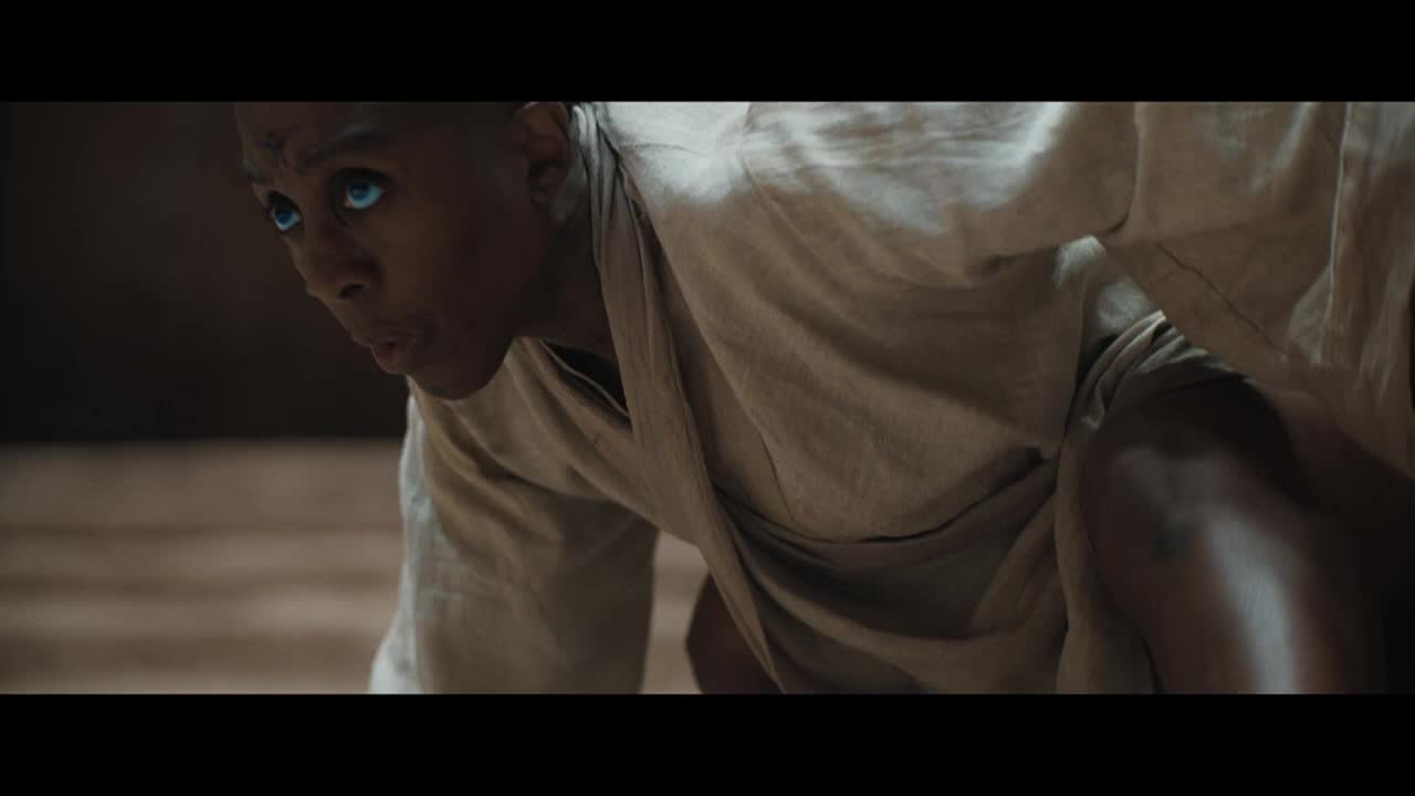 The person, clad in a light robe, is bent low, their intense blue eyes focused on something out of frame. Their posture suggests a moment of intense concentration, as if they are about to spring into action.
