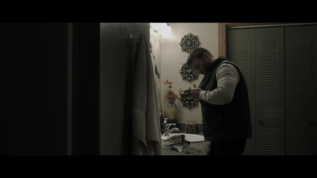 A man in a vest is buttoning it up in a bathroom, his reflection barely visible in the mirror. The room is dimly lit, with a towel hanging on the wall and a sink cluttered with items.
