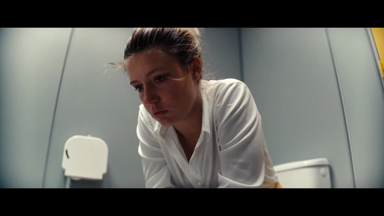 A young woman in a white shirt is hunched over, looking down at the toilet. Her face is etched with a mix of worry and sadness.
