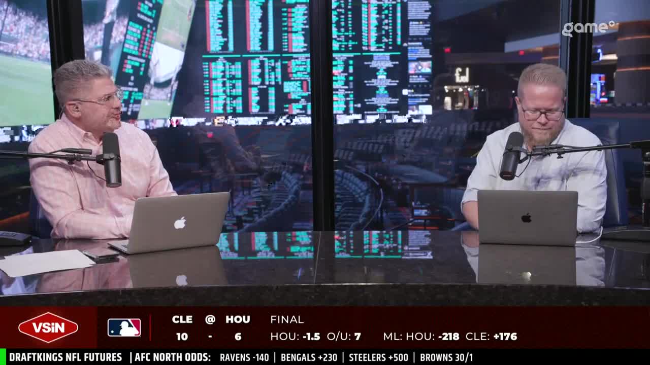 Two men sit behind a dark countertop, each with a laptop and microphone in front of them. The screen behind them displays a list of numbers and information, likely related to sports.
