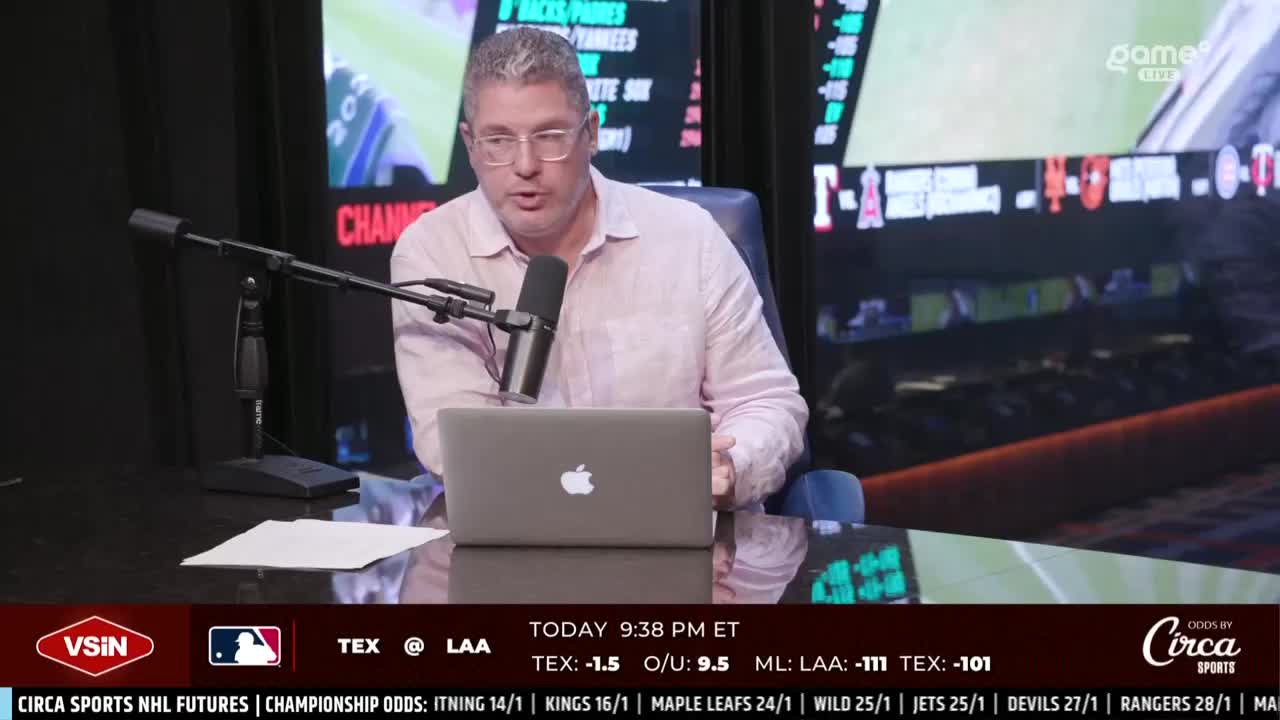 A man with glasses, wearing a light pink shirt, speaks into a microphone while looking at a laptop. Behind him, a screen displays sports scores and the "Game+" logo.
