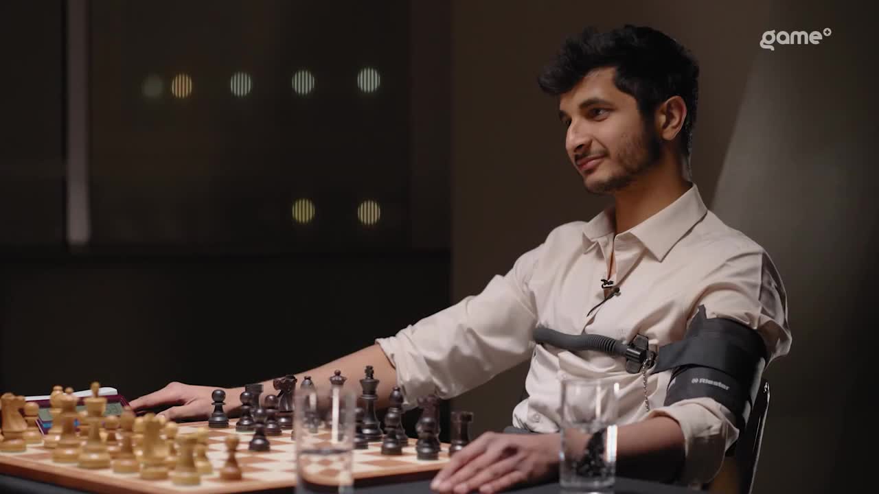 A young man sits at a chessboard, a sensor strapped to his arm. He's smiling slightly, gazing at the board as he considers his next move.
