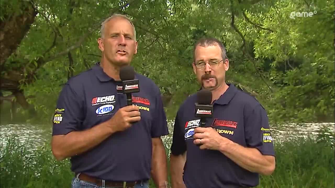 Two men in matching blue shirts and microphones stand in front of a backdrop of trees and water. They are speaking into the microphones, likely providing commentary for a Game+ broadcast from Canada.
