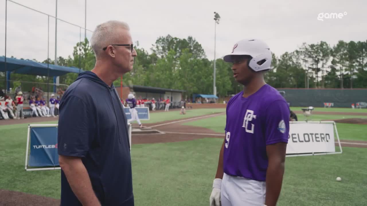 A coach, wearing a navy blue hoodie, is talking intently to a player in a purple jersey, both standing on the baseball diamond. The player, in a helmet, is listening, while other players and the dugout are in the background.
