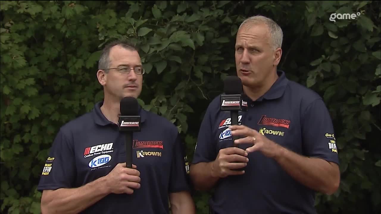 Two men in matching blue shirts, each holding a microphone, are standing in front of a wall of green foliage. The man on the right gestures with his hands as he speaks, while the other man listens intently.
