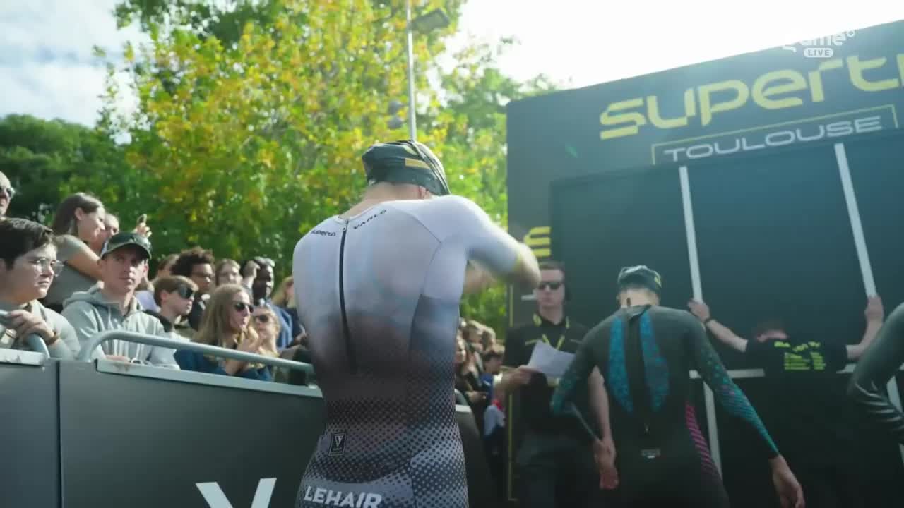 A competitor in a white and gray suit is adjusting his gear, facing the starting line. Spectators watch from behind a barrier, while others in wetsuits prepare to enter the water.
