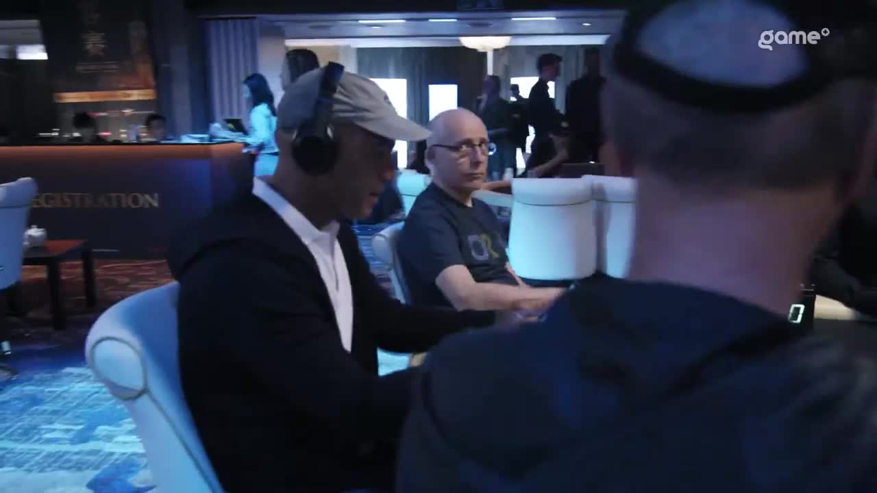 A man in a grey cap and headphones sits at a poker table, focused on the game. Across from him, another man with glasses stares intently, while the "game+" logo is visible in the corner.
