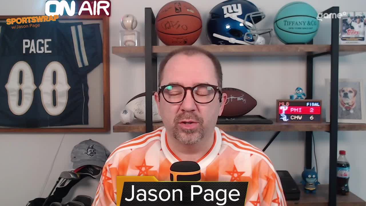 Jason Page, wearing glasses and an orange shirt, is speaking into a microphone. Behind him, a framed jersey and sports memorabilia are displayed on shelves, with a Game+ logo visible.
