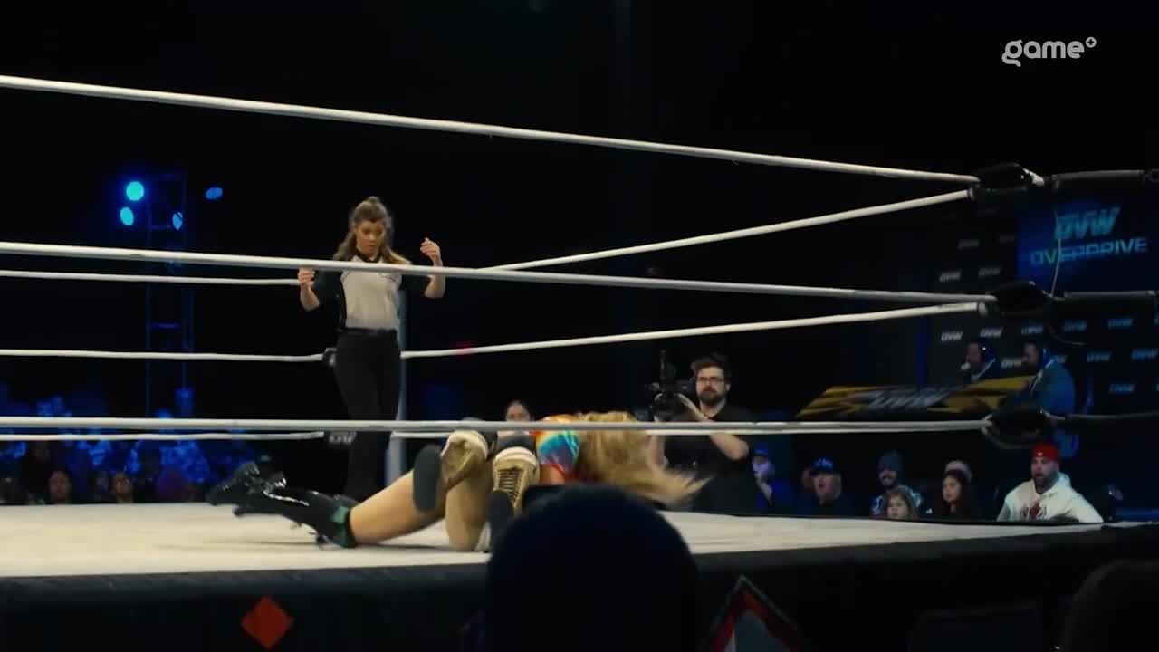 The wrestler lies on the mat, legs splayed, while the referee stands over her, arms raised. The crowd watches from ringside as the match unfolds on Game+.
