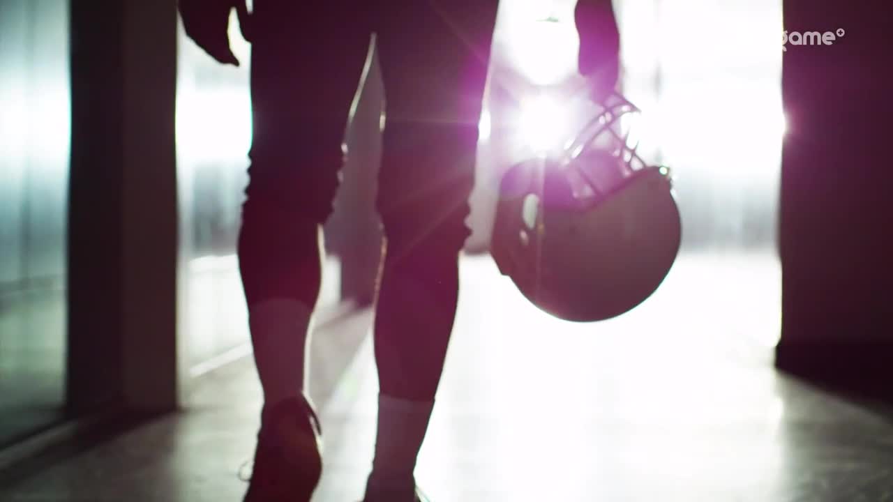 A football player, silhouetted against the bright light, walks away from me, helmet in hand. The player's legs are clad in maroon tights and white socks.

